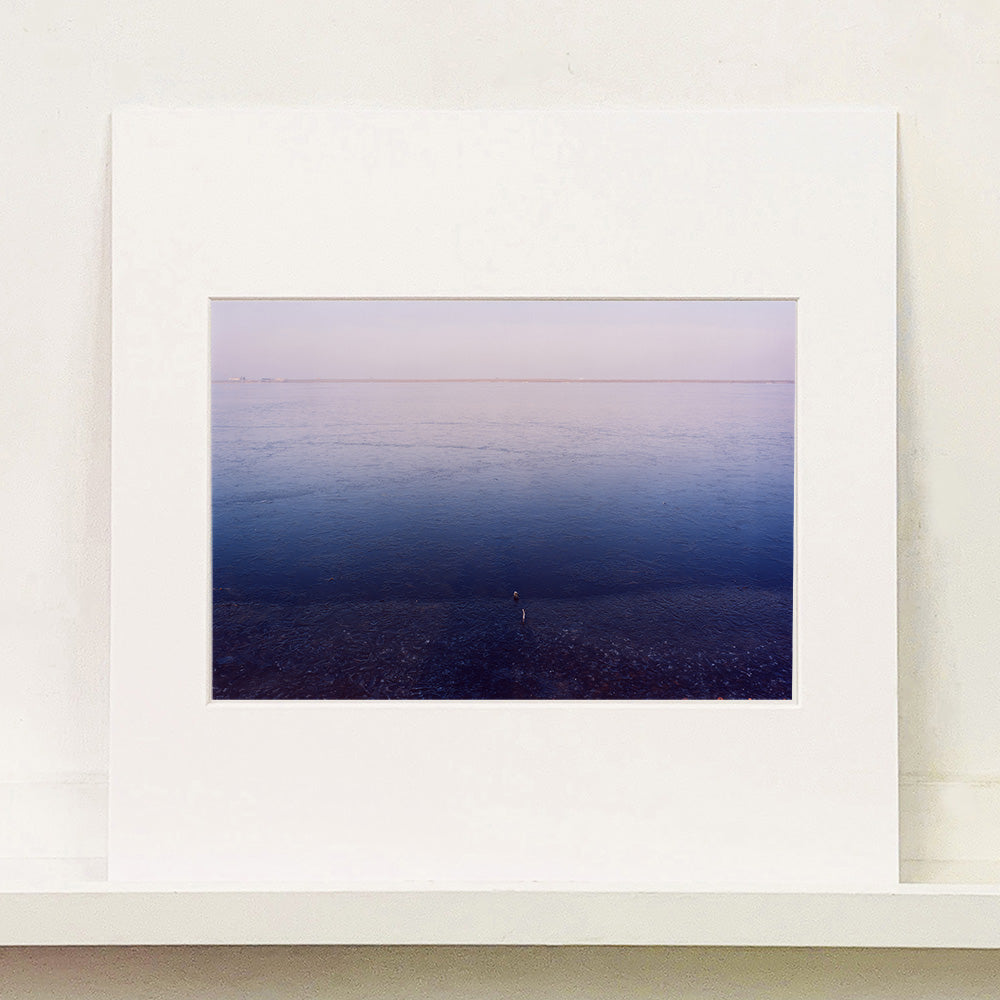 Mounted photograph by Richard Heeps. This photograph is looking towards the water. From a distance you can see two blocks of colour, blue at the bottom and lilac at the top, closer up you see the move of the water and a thin land strip along the horizon.