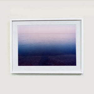 White framed photograph by Richard Heeps. This photograph is looking towards the water. From a distance you can see two blocks of colour, blue at the bottom and lilac at the top, closer up you see the move of the water and a thin land strip along the horizon.