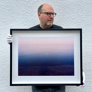 Black framed photograph held by photographer Richard Heeps. The held photograph is looking towards the water. From a distance you can see two blocks of colour, blue at the bottom and lilac at the top, closer up you see the move of the water and a thin land strip along the horizon.