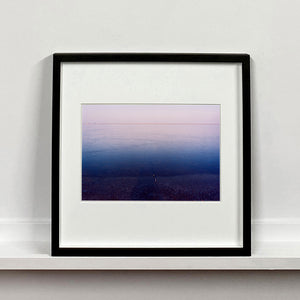Black framed photograph by Richard Heeps. This photograph is looking towards the water. From a distance you can see two blocks of colour, blue at the bottom and lilac at the top, closer up you see the move of the water and a thin land strip along the horizon.