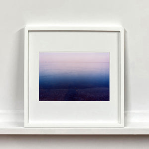 White framed photograph by Richard Heeps. This photograph is looking towards the water. From a distance you can see two blocks of colour, blue at the bottom and lilac at the top, closer up you see the move of the water and a thin land strip along the horizon.