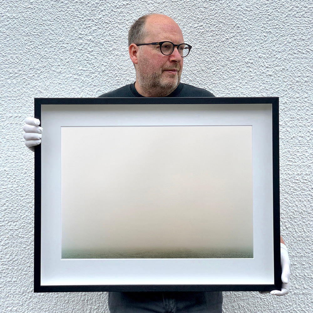 Black framed photograph held by photographer Richard Heeps. The photograph is of a wonderfully thick fog and all you can make out is the dark grey of the fenland  along the bottom of the photo.