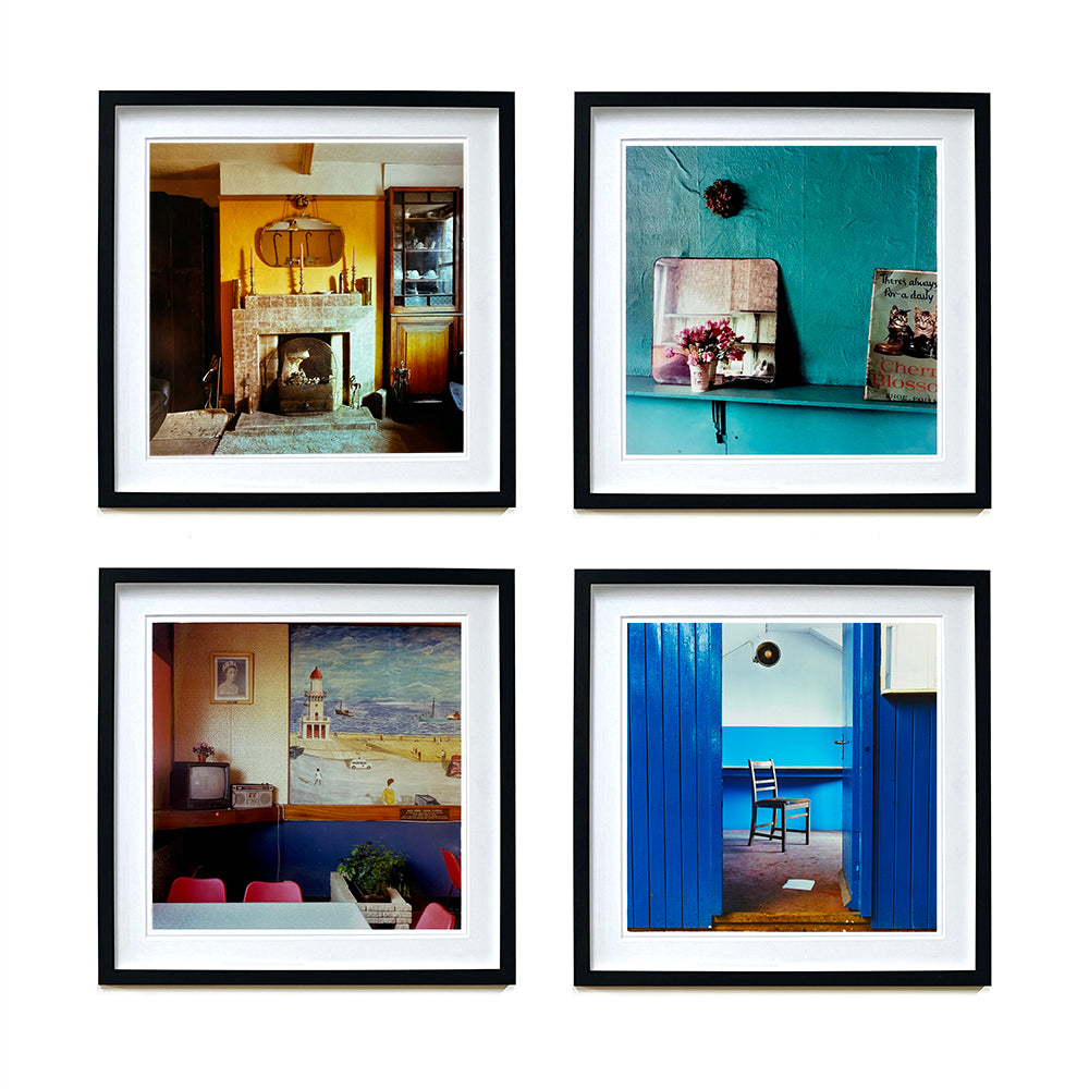 Four black framed photographs by Richard Heeps. From Richard's series Ordinary Places, the four photos capture vintage indoor scenes. The first one is a sitting room with a fireplace and candlesticks, the second a shelf with a mirror and a small bunch of flowers, the third is in the Fisherman's mission in the cafe with an old tv sitting in the corner, the fourth is through a blue door and you see a chair sitting by itself.