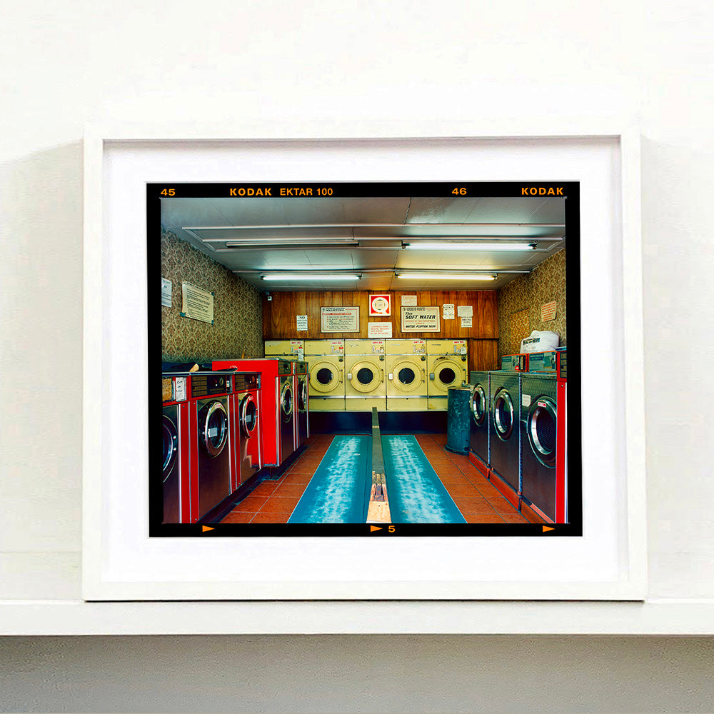 White framed photograph by Richard Heeps. A laundrette with washing machines on each wall and a double sided seat in the middle.