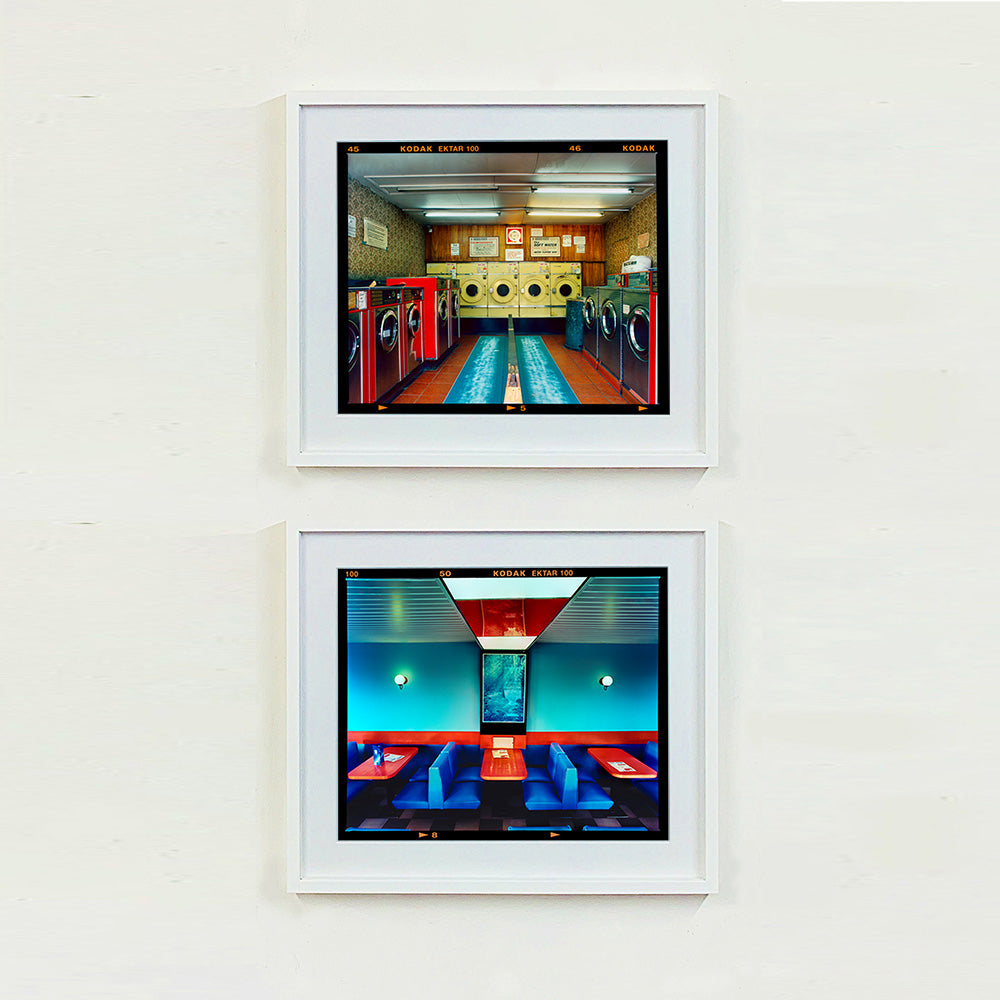 Two white framed photographs by Richard Heeps. The top one is of a laundrette with washing machines on each wall and a double sided seat in the middle. The bottom photograph is the inside of a restaurant with blue double sided seats, a blue wall and red tables.