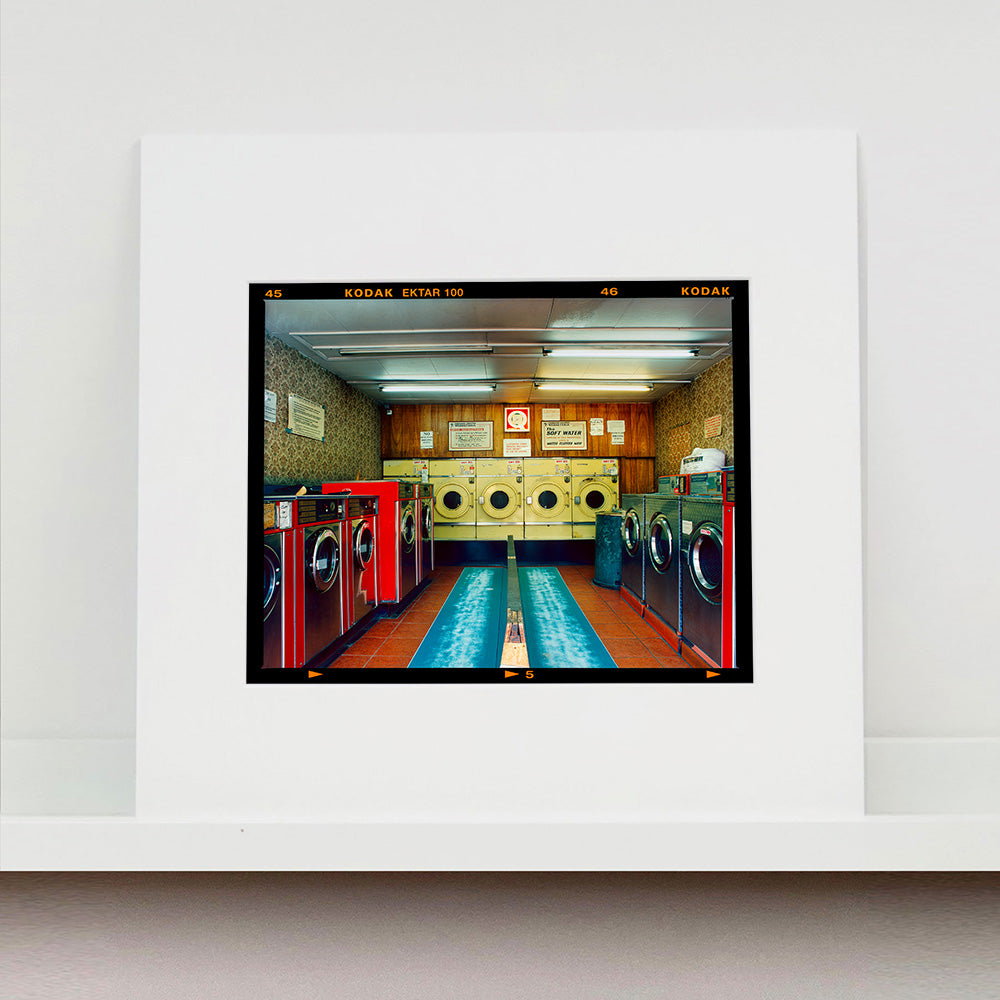 Mounted photograph by Richard Heeps. A laundrette with washing machines on each wall and a double sided seat in the middle.