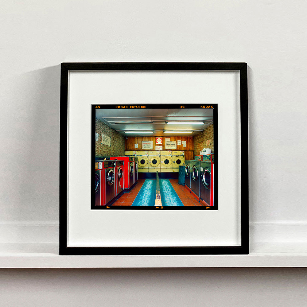 Black framed photograph by Richard Heeps. A laundrette with washing machines on each wall and a double sided seat in the middle.