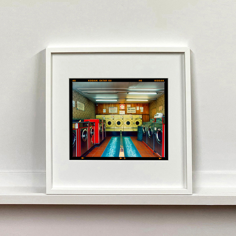 White framed photograph by Richard Heeps. A laundrette with washing machines on each wall and a double sided seat in the middle.