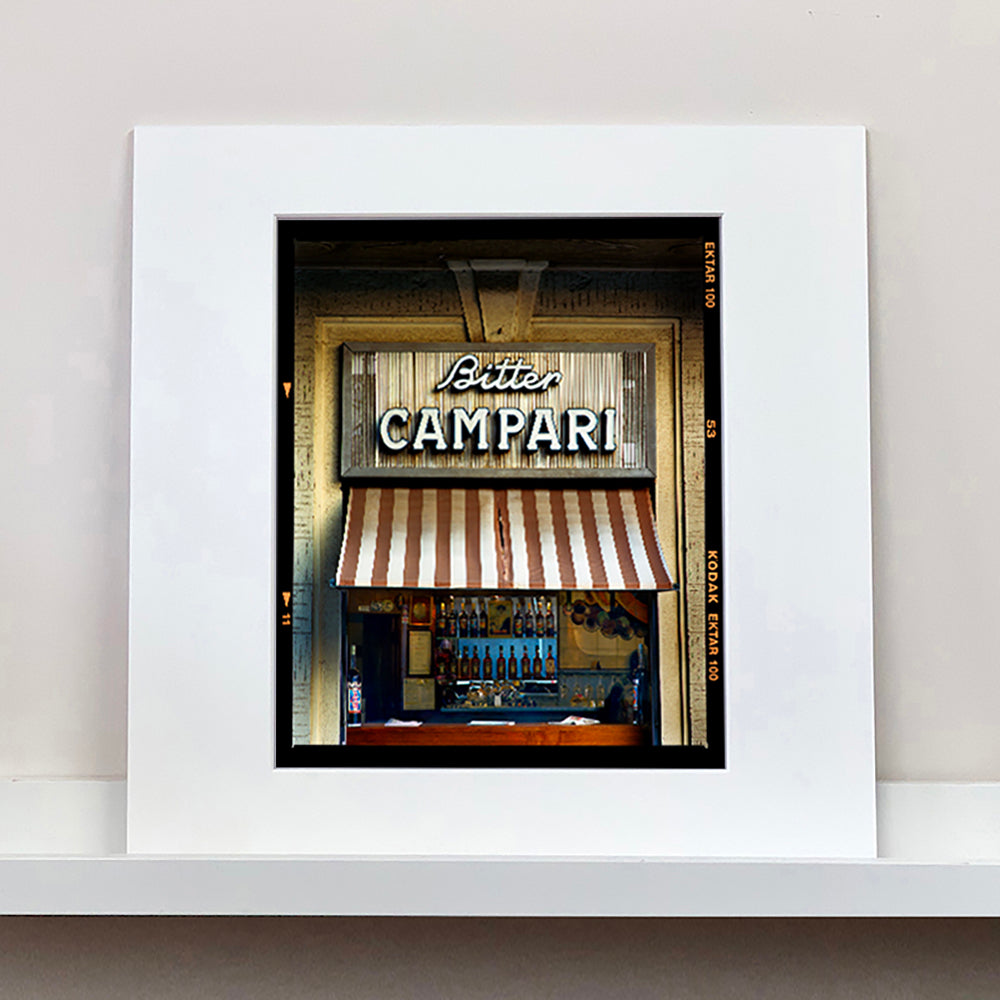 Photograph by Richard Heeps. A shop selling drinks, it has a red and white awning and the shop is called Bitter Campari in white lettering. This photograph has a black rebate.