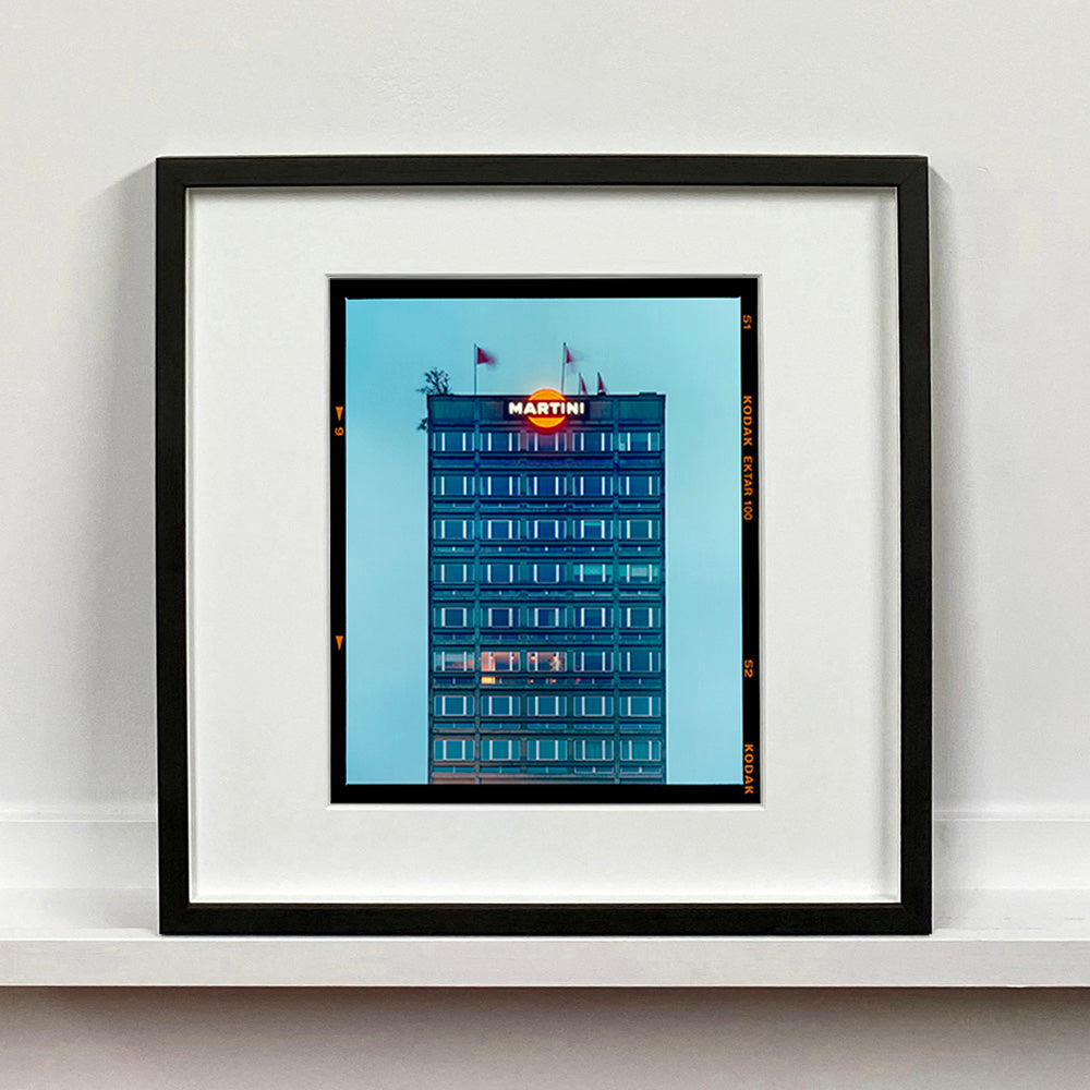 Black framed photograph by Richard Heeps. High rise offices in a blue light with Martini logo on the top facade. 