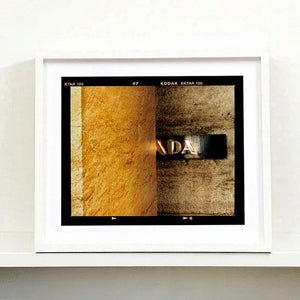 White framed photograph by Richard Heeps. Brown flecked marble walls in different tones. In the middle is half a brown plaque with golden letters showing half an A, followed by a D and an A.