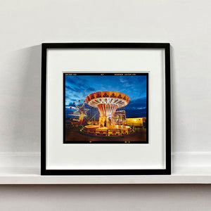Black framed photograph by Richard Heeps. A fairground ride, the chairoplanes, sits lit in golden and red colours against a dark blue sky.