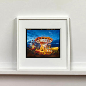 White framed photograph by Richard Heeps. A fairground ride, the chairoplanes, sits lit in golden and red colours against a dark blue sky.