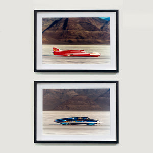 Two black framed photographs by Richard Heeps.  The top photograph is of a Red Ferguson Racing Streamliner sitting on a smooth salt flat with mountains in the background. The bottom photograph is of a Blue LSR Streamliner sitting on a smooth salt flat with mountains in the background.