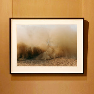 Black framed mounted photograph by Richard Heeps. A vague outline of the back of a light blue Buick car moving and obscured by the dust clouds which it has created.