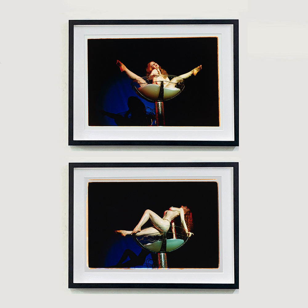 Two black framed photographs by Richard Heeps. In the top photograph, a burlesque dancer sits in a human sized champagne glass. The glass is filled with a green frothy liquid and in the middle sits the dancer with her legs sticking out over the rim of the glass. The bottom photograph has the dancer balancing on the top of the champagne glass, with her body arched up.