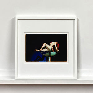 White framed photograph by Richard Heeps. Burlesque Dancer arches her back while balancing over a human size champagne glass.