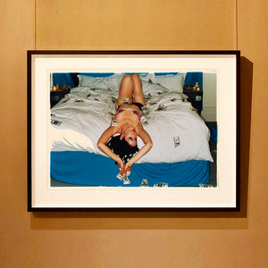 Black framed photograph by Richard Heeps. A woman lies semi naked on a bed with money surrounding her. The photograph is taken from the end of the bed so the woman appears upside down. The bed has a white duvet with blue under sheets.