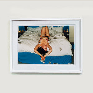 White framed photograph by Richard Heeps. A woman lies semi naked on a bed with money surrounding her. The photograph is taken from the end of the bed so the woman appears upside down. The bed has a white duvet with blue under sheets.