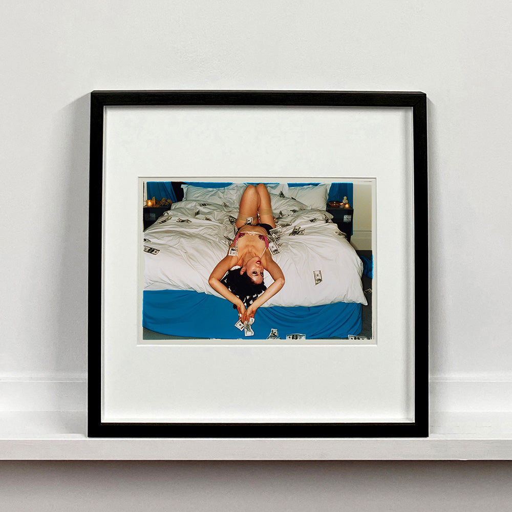 Black framed photograph by Richard Heeps. A woman lies semi naked on a bed with money surrounding her. The photograph is taken from the end of the bed so the woman appears upside down. The bed has a white duvet with blue under sheets.