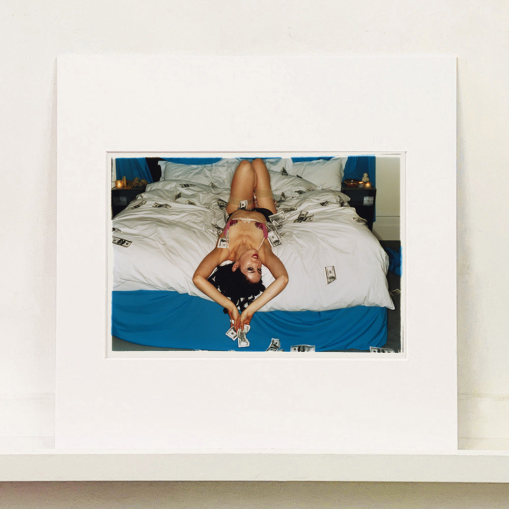 Mounted photograph by Richard Heeps. A woman lies semi naked on a bed with money surrounding her. The photograph is taken from the end of the bed so the woman appears upside down. The bed has a white duvet with blue under sheets.