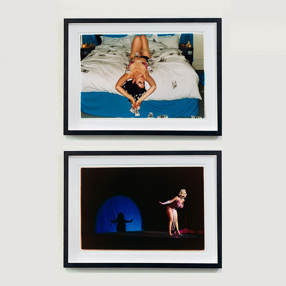 Two black framed photographs by Richard Heeps. The top one is of a woman who lies semi naked on a bed with money surrounding her. The photograph is taken from the end of the bed so the woman appears upside down. The bed has a white duvet with blue under sheets. The bottom photograph has a burlesque dancer taking a bow on a dark stage. The only other light is a blue half circle of light which reflects her silhouette. 