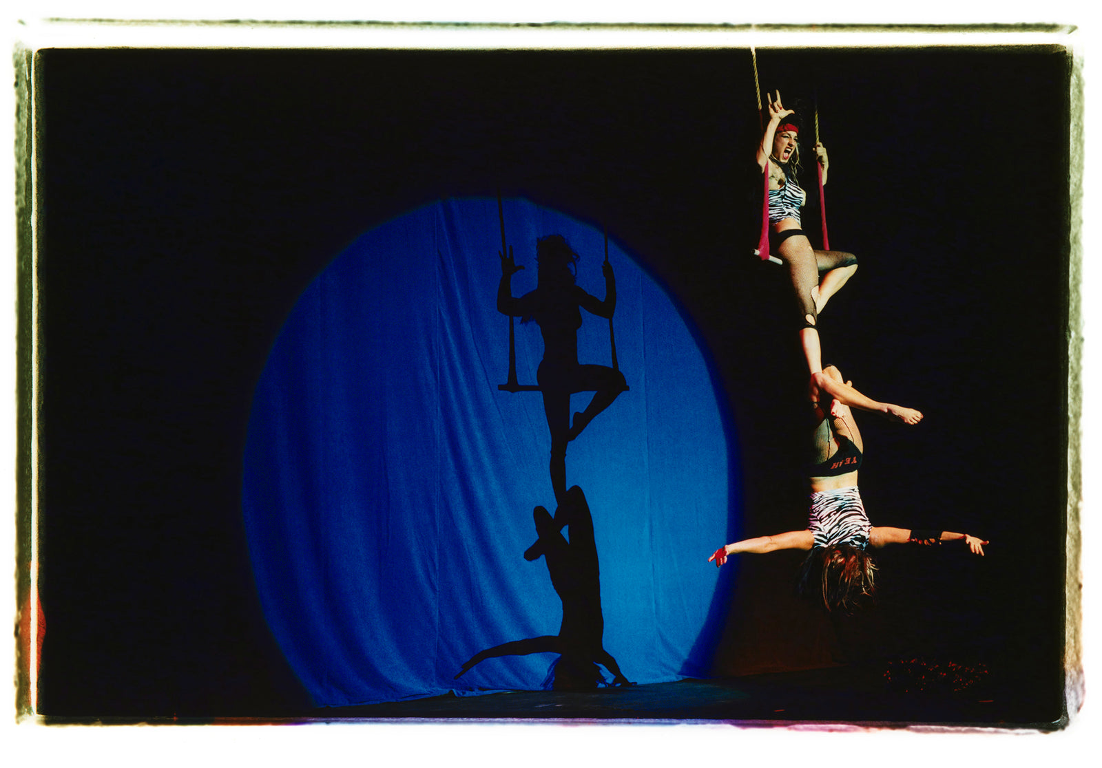 Photograph by Richard Heeps. The photo is of two women on a trapeze, one sits on the swing and the other is hanging down from the first. The background is dark apart from a blue circle in which shows their acrobatic shadow.