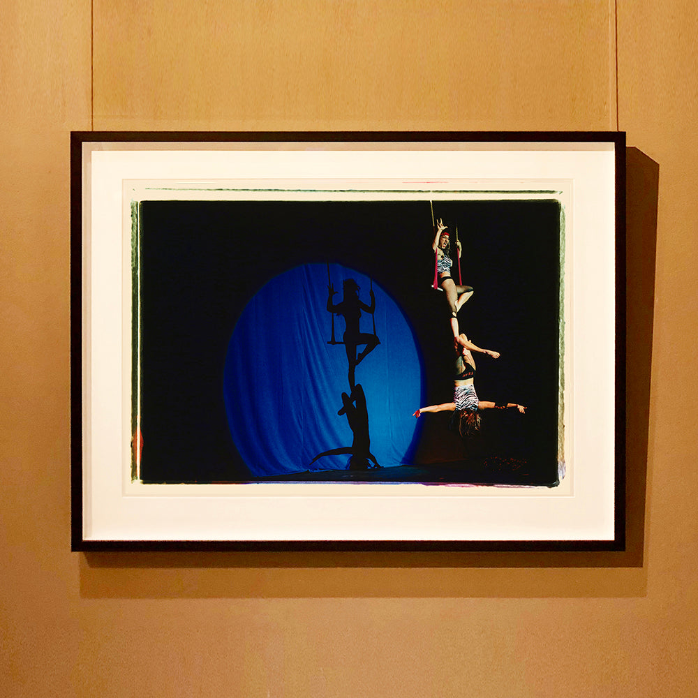 Black framed photograph by Richard Heeps. The photo is of two women on a trapeze, one sits on the swing and the other is hanging down from the first. The background is dark apart from a blue circle in which shows their acrobatic shadow.