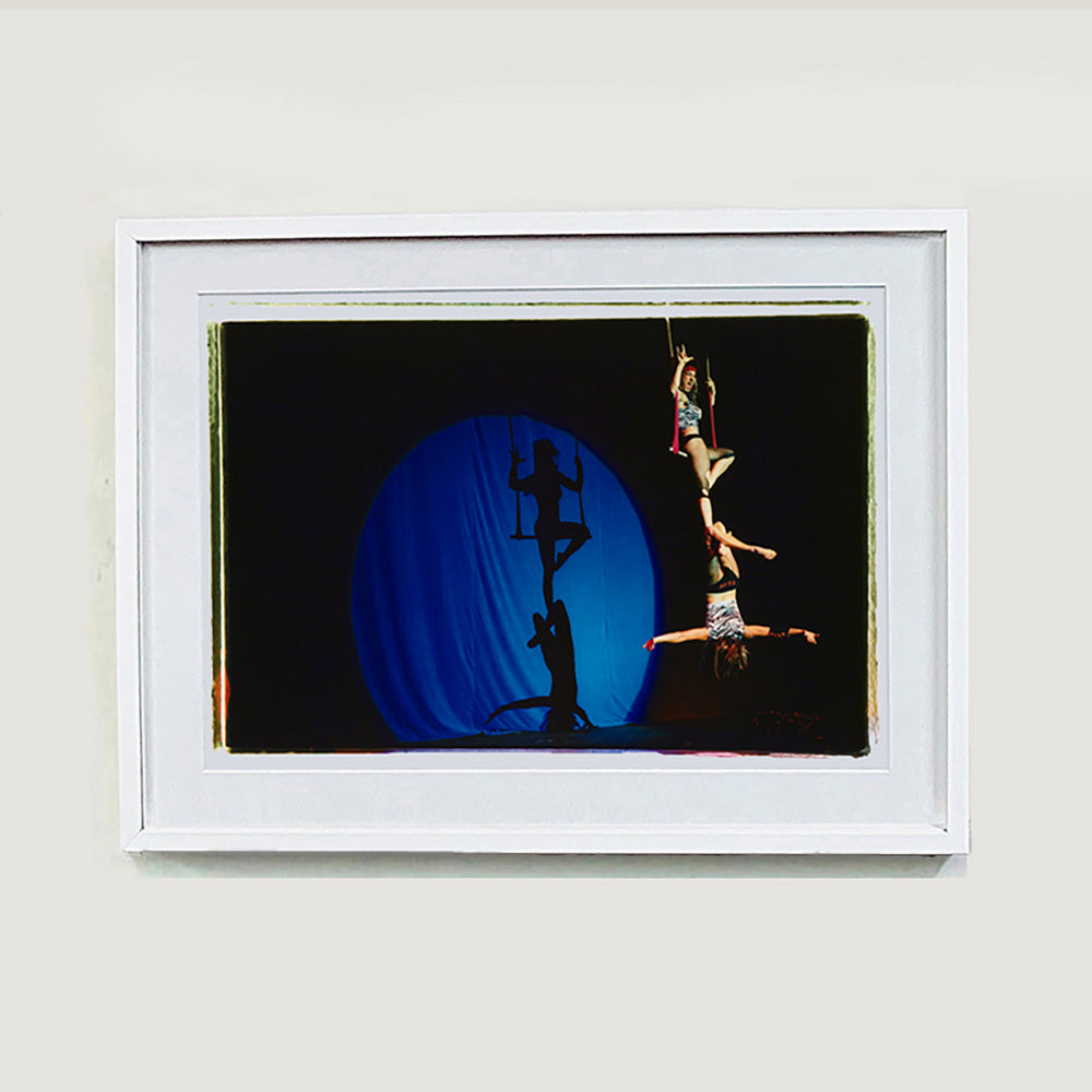 White framed photograph by Richard Heeps. The photo is of two women on a trapeze, one sits on the swing and the other is hanging down from the first. The background is dark apart from a blue circle in which shows their acrobatic shadow.