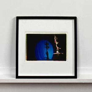 Black framed photograph by Richard Heeps. The photo is of two women on a trapeze, one sits on the swing and the other is hanging down from the first. The background is dark apart from a blue circle in which shows their acrobatic shadow.