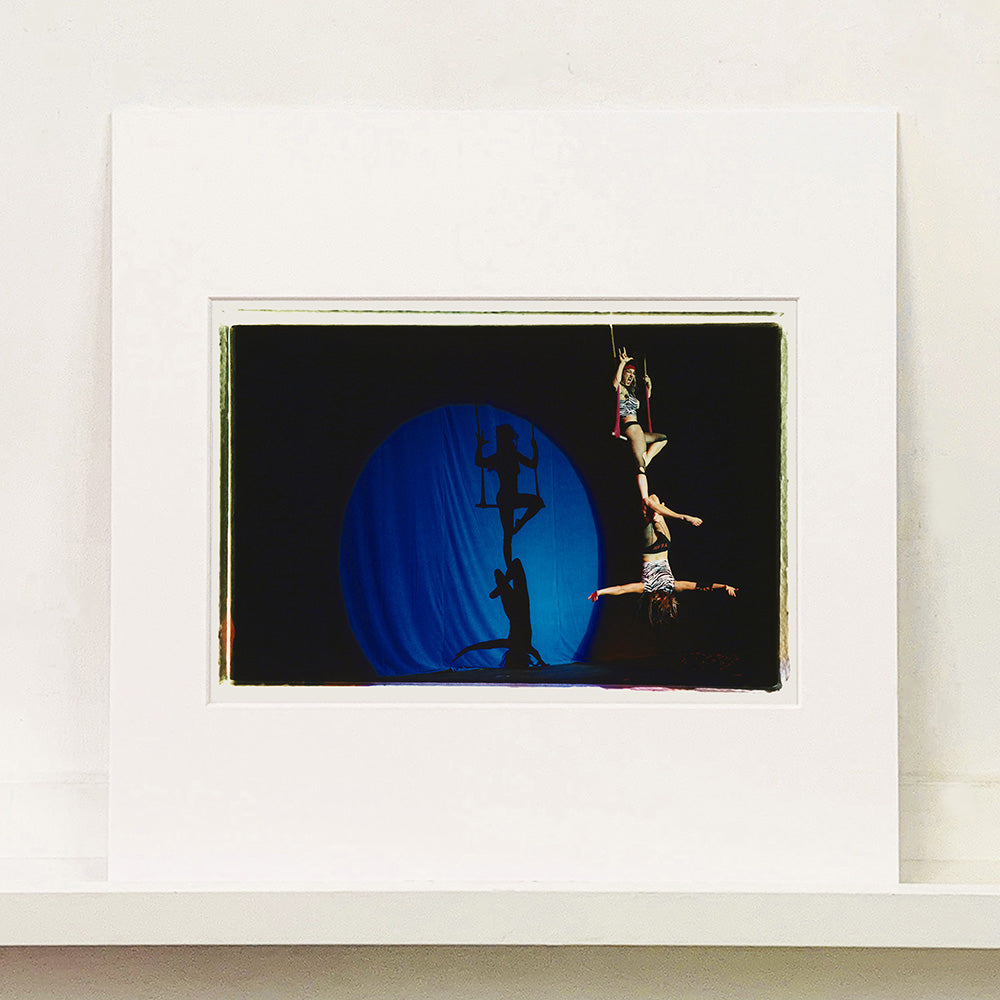 Mounted photograph by Richard Heeps. The photo is of two women on a trapeze, one sits on the swing and the other is hanging down from the first. The background is dark apart from a blue circle in which shows their acrobatic shadow.