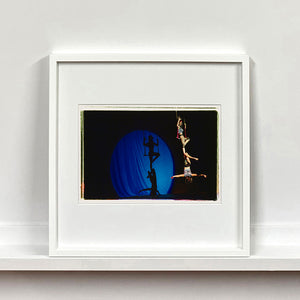 White framed photograph by Richard Heeps. The photo is of two women on a trapeze, one sits on the swing and the other is hanging down from the first. The background is dark apart from a blue circle in which shows their acrobatic shadow.
