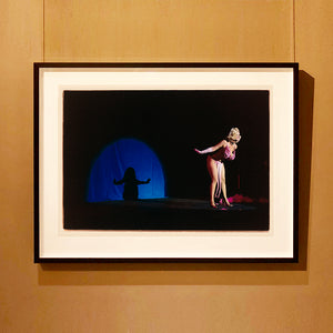 Black framed photograh by Richard Heeps. A Burlesque dancer with a pink fluffy bikini bows on a black stage, in the background is a blue circle containing her silhouette.