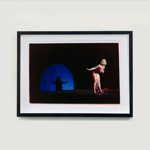Black framed photograh by Richard Heeps. A Burlesque dancer with a pink fluffy bikini bows on a black stage, in the background is a blue circle containing her silhoette.