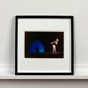 Black framed photograh by Richard Heeps. A Burlesque dancer with a pink fluffy bikini bows on a black stage, in the background is a blue circle containing her silhouette.