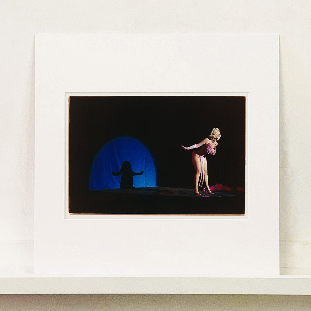 Mounted photograh by Richard Heeps. A Burlesque dancer with a pink fluffy bikini bows on a black stage, in the background is a blue circle containing her silhouette.