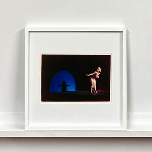 White framed photograh by Richard Heeps. A Burlesque dancer with a pink fluffy bikini bows on a black stage, in the background is a blue circle containing her silhouette.
