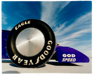 Photograph by Richard Heeps. This photograph has the tyre and the very front tip of a drag car. The car's name is written on the front end "God Speed". Behind the car are white vertical clouds shooting through a blue sky.