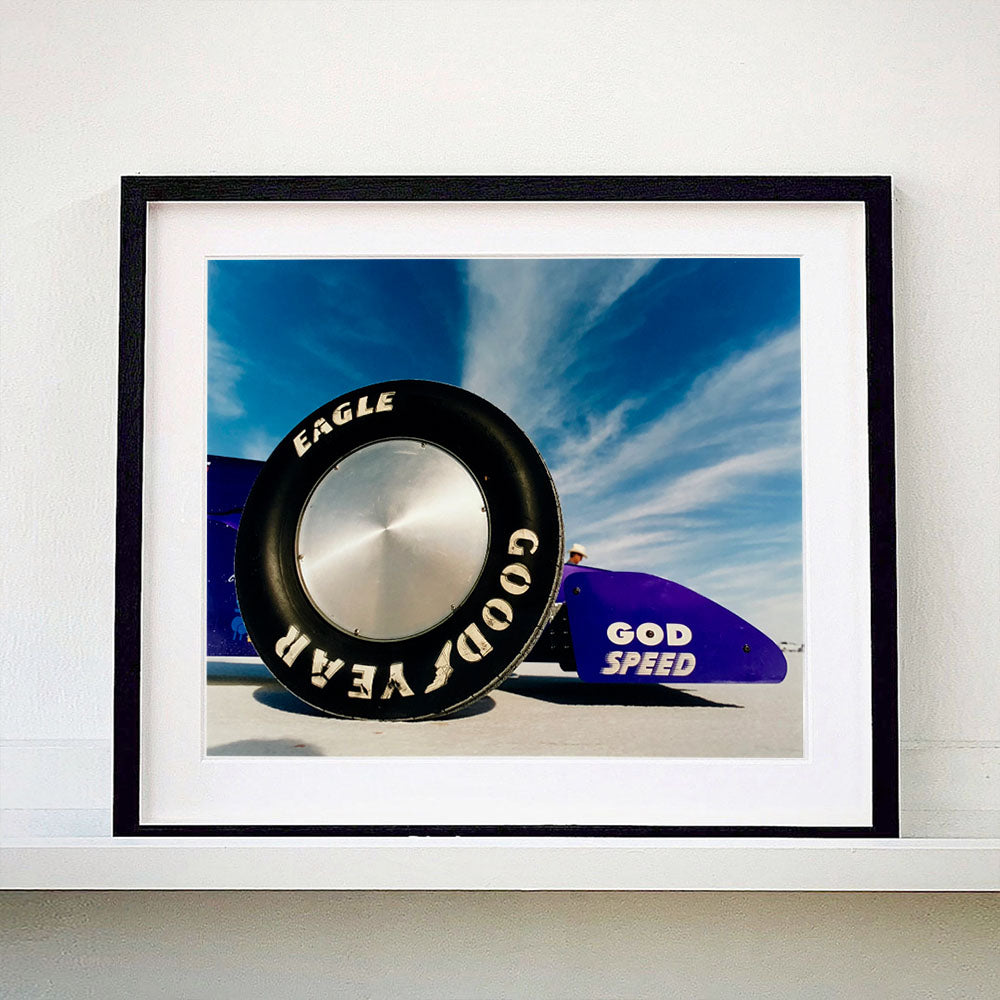 Black framed photograph by Richard Heeps. This photograph has the tyre and the very front tip of a drag car. The car's name is written on the front end "God Speed". Behind the car are white vertical clouds shooting through a blue sky.