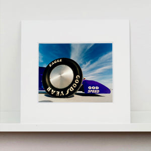 Mounted photograph by Richard Heeps. This photograph has the tyre and the very front tip of a drag car. The car's name is written on the front end "God Speed". Behind the car are white vertical clouds shooting through a blue sky.