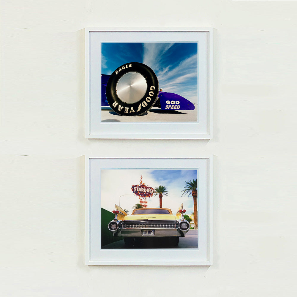 Two white framed photographs by Richard Heeps. The top photograph has the tyre and the very front tip of a drag car. The car's name is written on the front end "God Speed". Behind the car are white vertical clouds shooting through a blue sky. The bottom photo features Las Vegas and has the back of a yellow chevy car and above the car sits the sign for the Stardust casino.