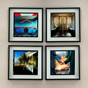 4 black framed photographs in a square by Richard Heeps. The first photograph captures the inside of a Wimpy Restaurant in Norfolk. There is bright blue seats and red tables. The walls are blue and there is a big red chevron light attached to the wall. The second photograph is the inside of a subway car in NY, the bottom left photograph is a lovely palm tree outside a cream building on the beach. The last photograph on the bottom right is two hands held with a fire roaring behind, and trees behind the fire.