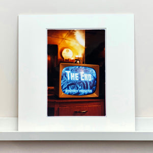 Mounted photograph by Richard Heeps. A retro small brown television sits on a wooden table displaying The End on a black and white film which shows as blue and white. In the background of the television is a warm light.