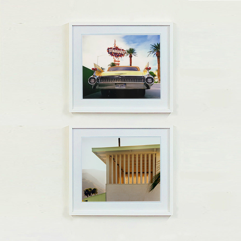 Two white framed photographs by Richard Heeps.  The top photograph features the back end of the classic American car with a number place DREAM01 sits underneath the STARDUST casino sign. The photo at the bottom features a cream building with a big window with vertical blinds, a palm frond appears from the right hand side and to the left of the building in the distance are misty covered mountains and trees.