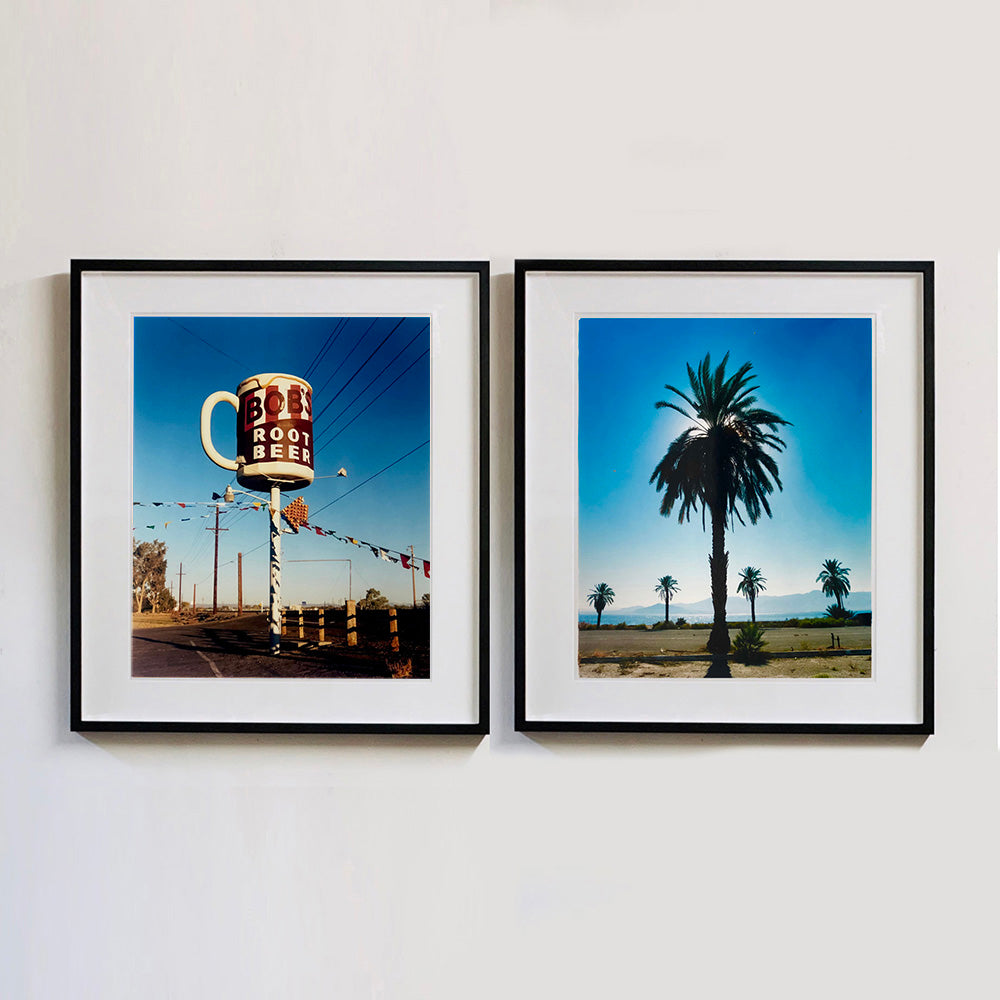 Two black framed photographs by Richard Heeps. A giant model of a mug with Bob's Root Beer written on it sits on top of a giant pole. There is bunting hanging from the pole. It sits alongside a power line on a remote looking American country road. The photograph on the right hand side features a palm tree with palm trees in the background, set in front of a blue sky and the sun filtering from behind the fronds of the tree.
