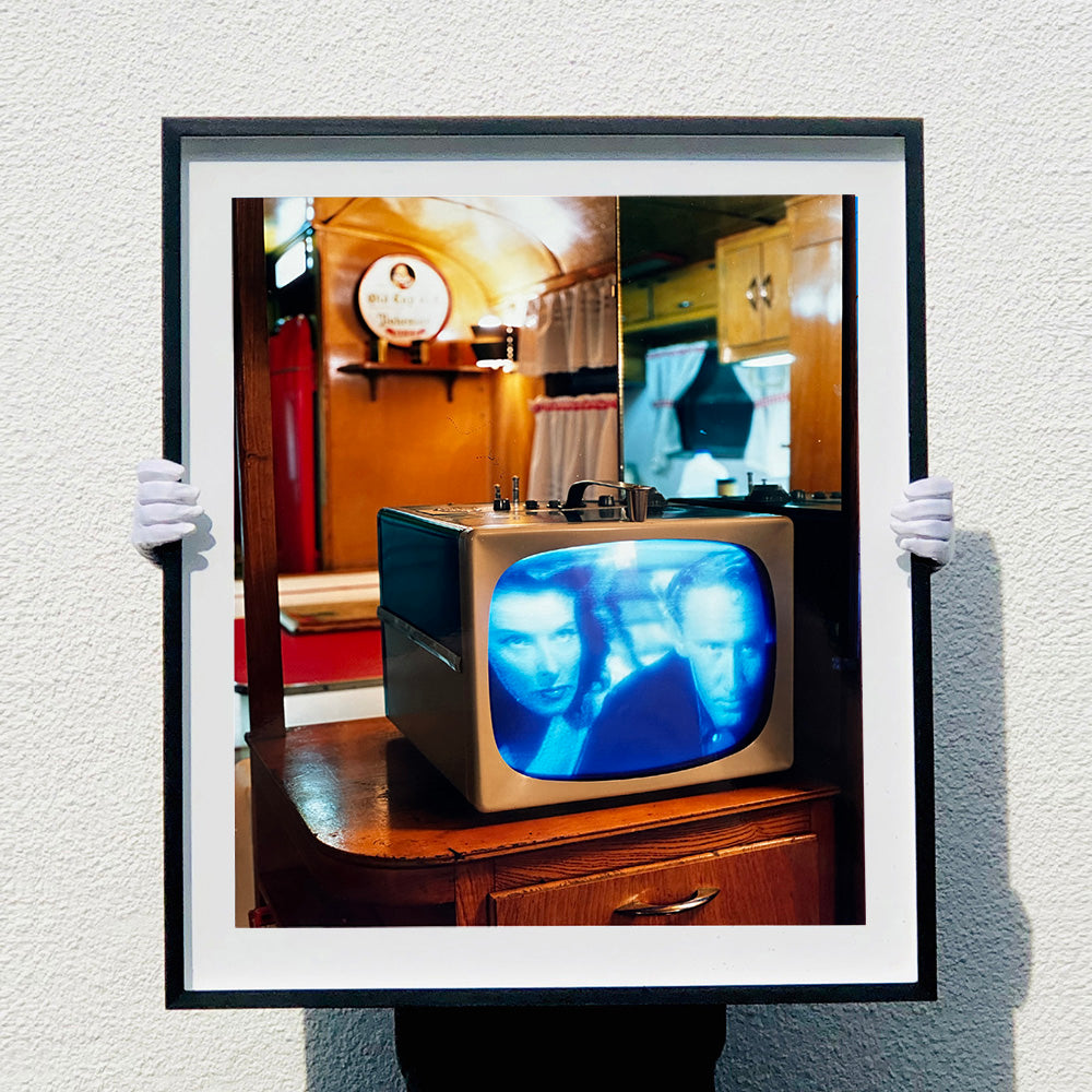 Black framed photograph by Richard Heeps. A retro small brown television sits on a wooden table displaying a scene from a black and white film, Without Love. The image shows as blue and white. In the background of the television is a warm wood-panelled light.