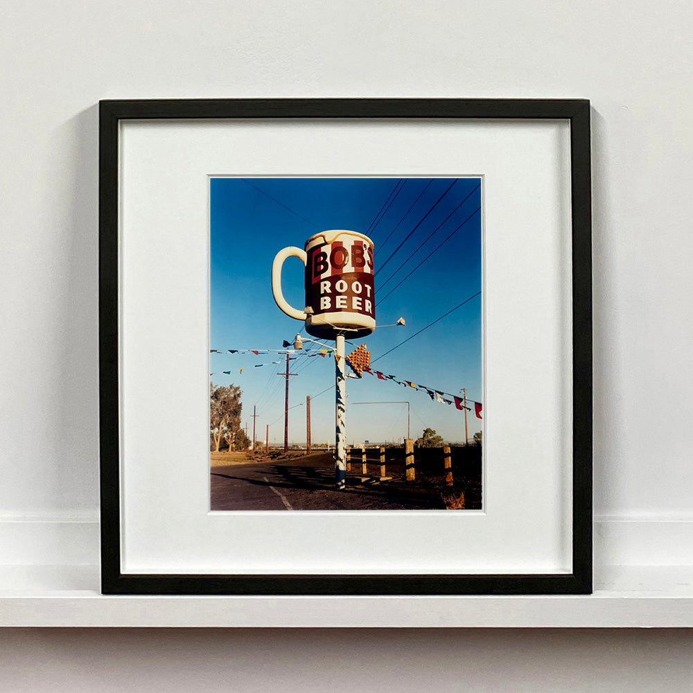 Black framed photograph by Richard Heeps. A giant model of a mug with Bob's Root Beer written on it sits on top of a giant pole. There is bunting hanging from the pole. It sits alongside a power line on a remote looking American country road.