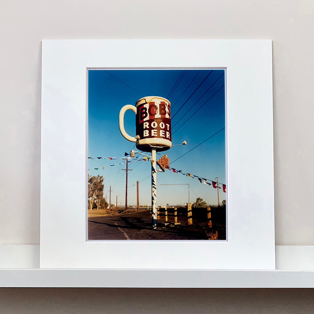 Mounted photograph by Richard Heeps. A giant model of a mug with Bob's Root Beer written on it sits on top of a giant pole. There is bunting hanging from the pole. It sits alongside a power line on a remote looking American country road.