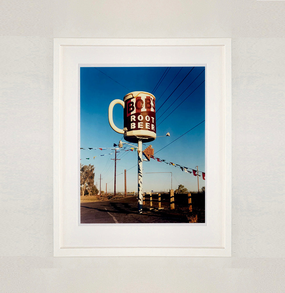 White framed photograph by Richard Heeps. A giant model of a mug with Bob's Root Beer written on it sits on top of a giant pole. There is bunting hanging from the pole. It sits alongside a power line on a remote looking American country road.