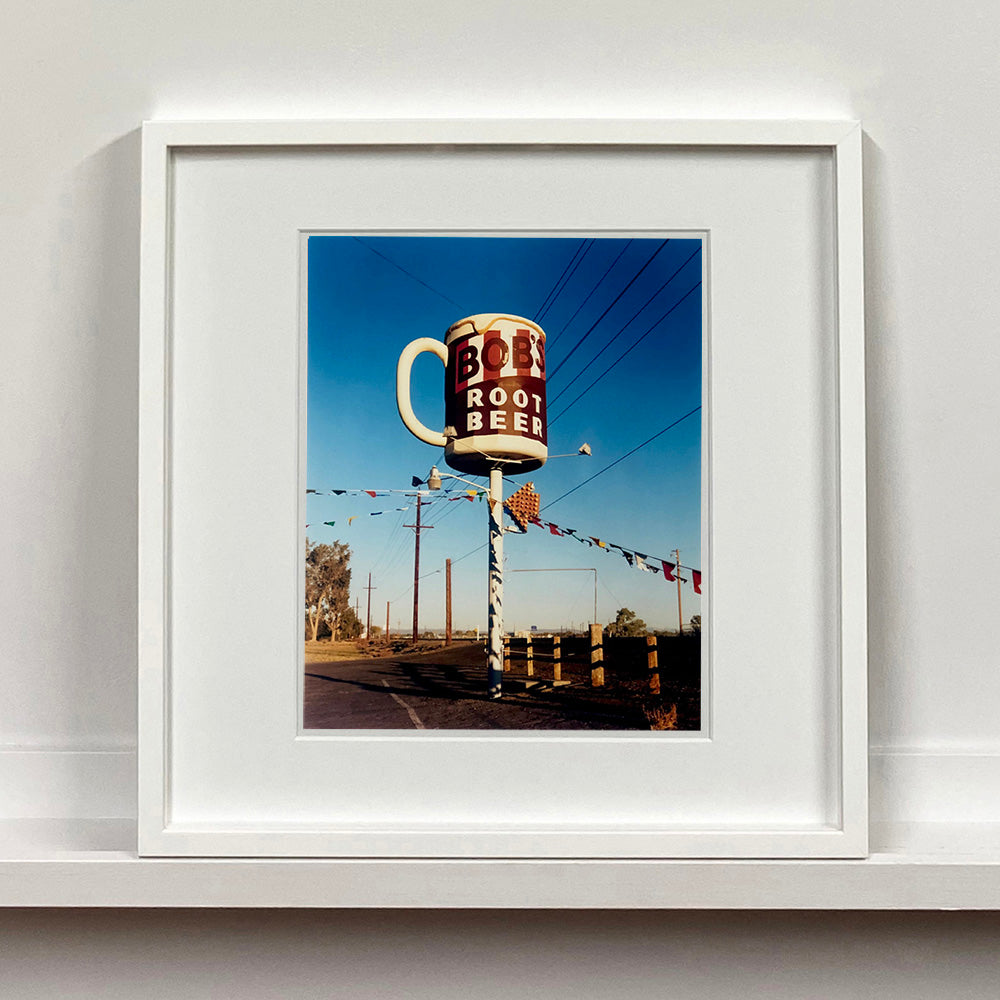 White framed photograph by Richard Heeps. A giant model of a mug with Bob's Root Beer written on it sits on top of a giant pole. There is bunting hanging from the pole. It sits alongside a power line on a remote looking American country road.